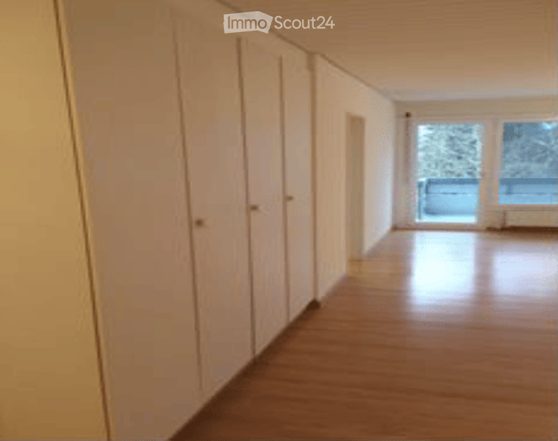 Empty room with wooden flooring and white walls, featuring several closed doors and large windows with a view of the outside.