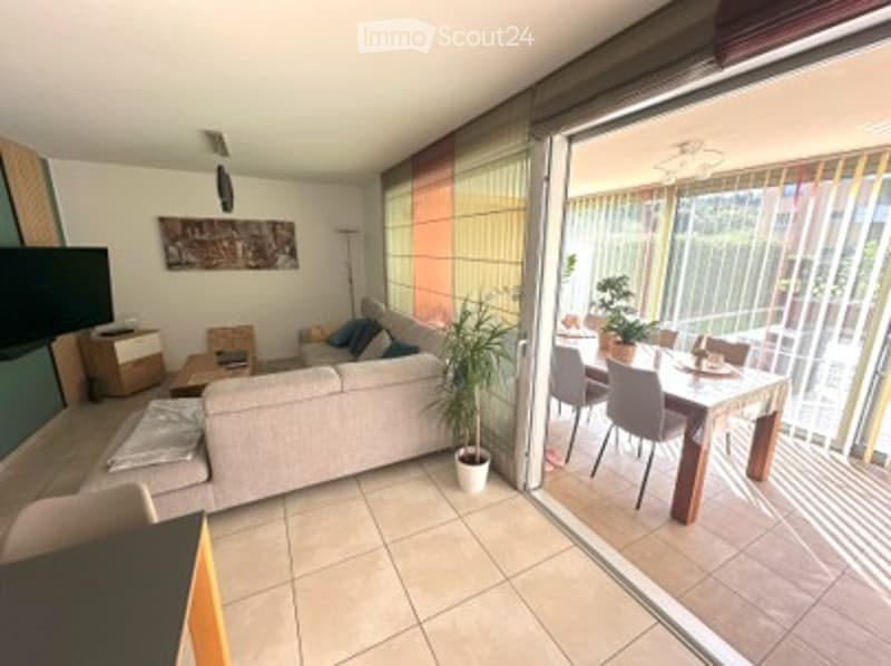 Bright living and dining area with large sliding glass doors opening to a balcony. Features a grey sofa, coffee table, dining table with chairs, and a potted plant.
