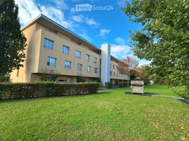 Modern multi-story building with a flat roof and rectangular windows, surrounded by a well-maintained lawn and trees.