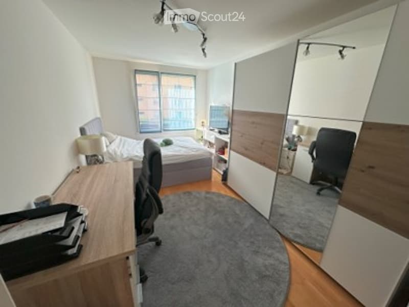 A modern, well-lit bedroom with a large bed, a desk with a chair, a round rug, and a large mirror on the wall. The room features contemporary furniture and a sleek design.