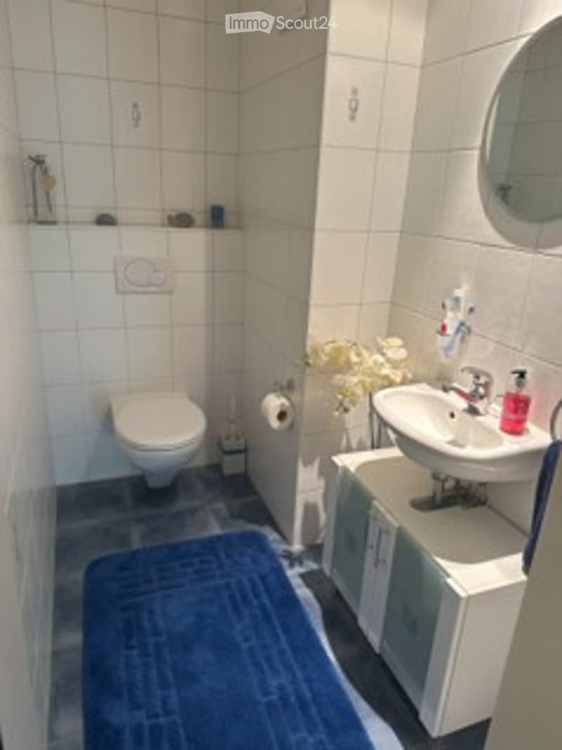 Modern bathroom with white tiled walls, a toilet, a sink with a cabinet, a mirror, and a blue bath rug on the floor.