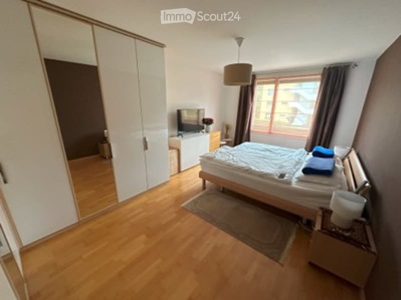 Bright bedroom with wooden flooring, large window with view, bed, wardrobe, TV, nightstand, and lamp.