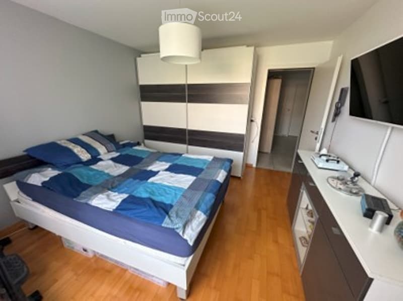 A modern, well-lit bedroom with a large bed, a wardrobe with a mirror, a sink, and a lamp. The room features wooden flooring and a neutral color palette.
