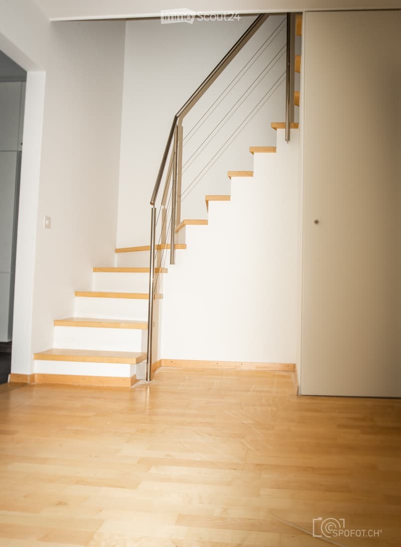 Modern staircase with wooden steps and a metal railing, leading up from a wooden floor hallway.
