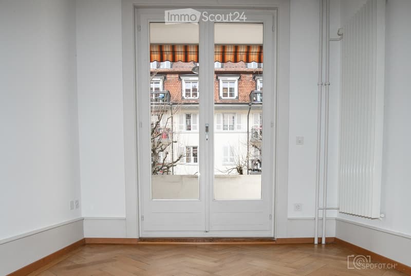 Empty room with wooden flooring, white walls, and a large white sliding door leading to a view of neighboring buildings.