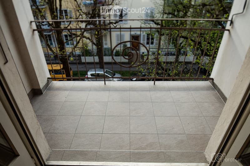 Balcony with tiled floor and metal railing, overlooking a street with parked cars and trees.