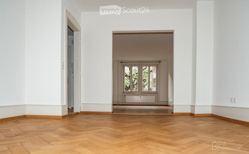 Empty room with wooden flooring, white walls, and large windows in the background.