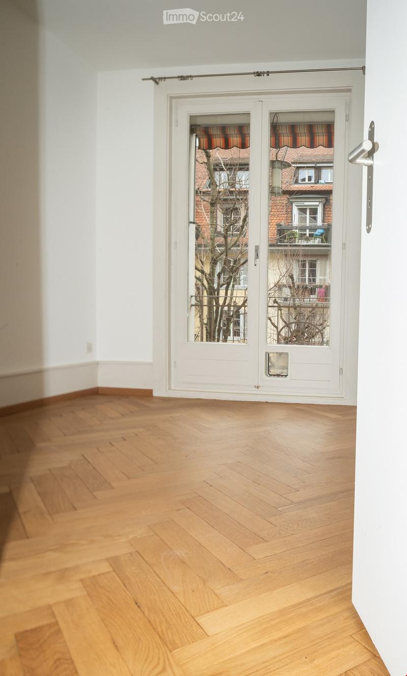 Empty room with wooden herringbone flooring, white walls, and a large white sliding door leading to a balcony with a view of neighboring buildings and a tree.