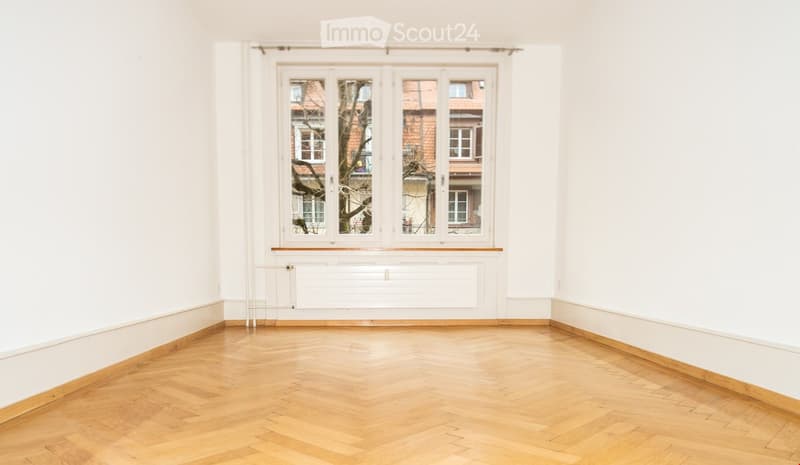 Empty room with wooden floor, white walls, large window with white frame, radiator below window, and baseboard trim.