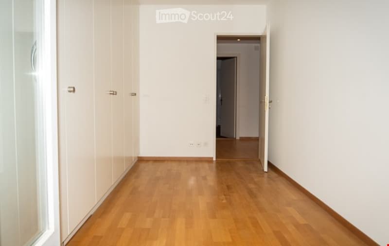 Empty hallway with light wooden flooring and white walls, featuring built-in white closet doors on one side and an open doorway leading to another room.