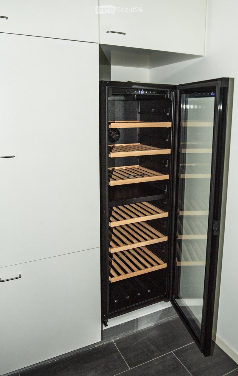 A modern wine cooler with multiple wooden shelves, integrated into a sleek kitchen with white cabinets and dark tiled flooring.
