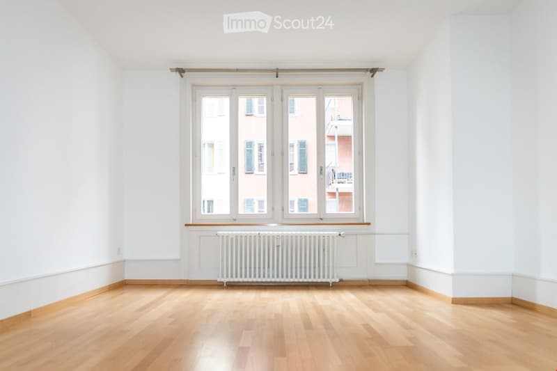 Empty room with light wooden flooring, white walls, large windows with shutters, and a radiator below the windows.