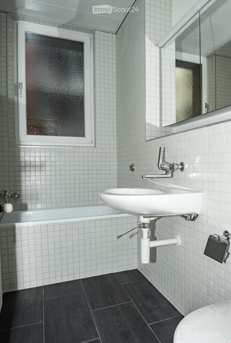 Modern bathroom with white tiled walls, dark tiled floor, white sink, bathtub, and large mirror.