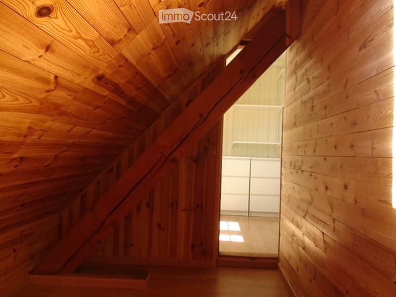A narrow wooden hallway with exposed wooden beams and a slanted ceiling, leading to a bright, open space with white walls and a glass door.
