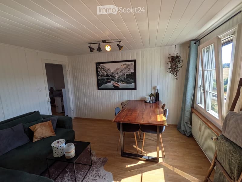 Cozy living room with a green sofa, coffee table, dining table, and chairs. The room features wooden flooring, large windows with curtains, and a picture frame on the wall.
