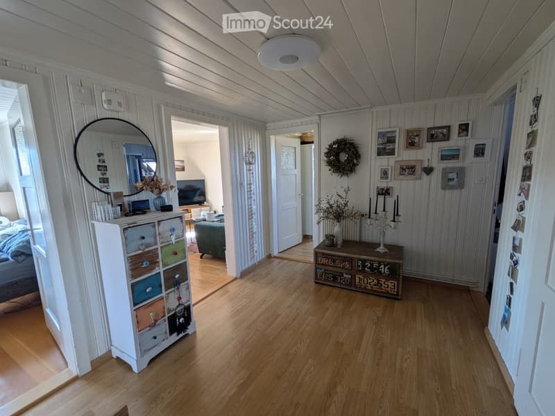 Cozy hallway with wooden flooring, white walls, and various decorative items including a mirror, framed photos, and a small table with candles and a wreath.