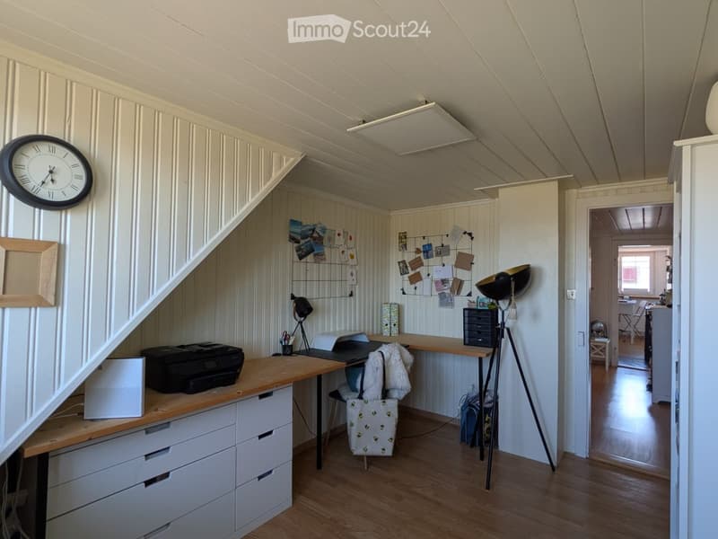 A well-lit, modern home office with a wooden desk, laptop, printer, and a large clock on the wall. The room features white paneled walls, a slanted ceiling, and a cozy chair with a fur throw.