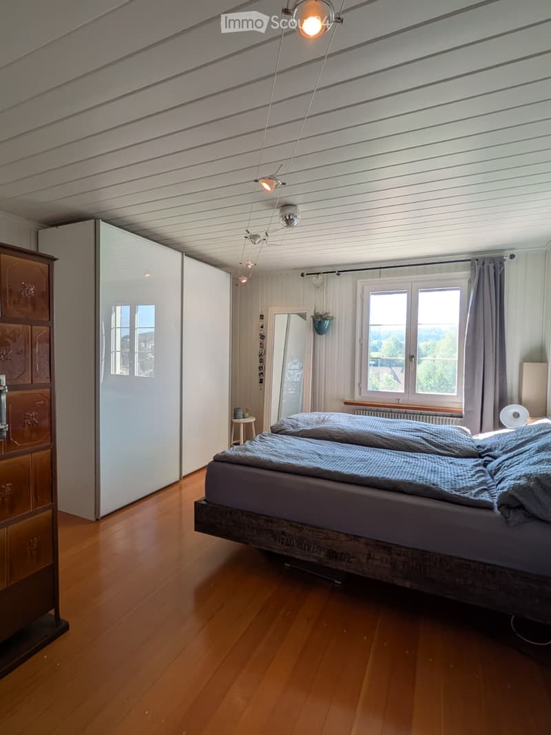 Bright bedroom with wooden flooring, a large bed with grey bedding, a wardrobe, a small stool, a mirror, and a window with curtains.