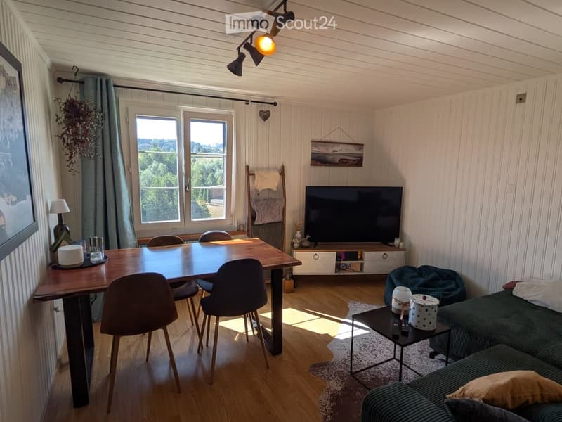 Bright living room with wooden flooring, large windows, a green sofa, coffee table, dining table with chairs, and a TV stand.