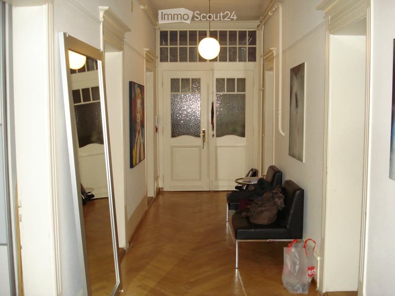 A hallway with wooden flooring, white walls, and a hanging light fixture. A small seating area with a sofa, table, and chair is visible, along with a handbag and shopping bag on the floor.
