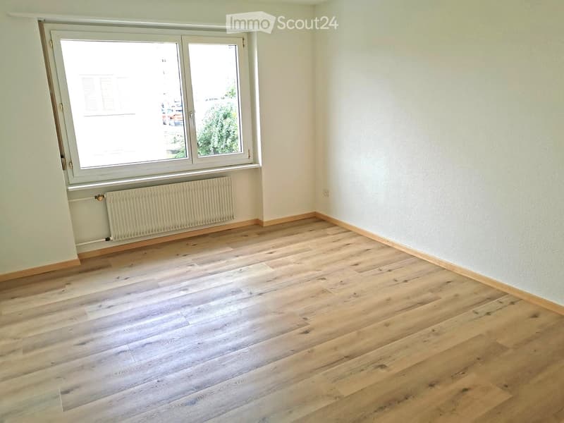 Empty room with light wooden flooring, white walls, and a large window with a radiator below it.
