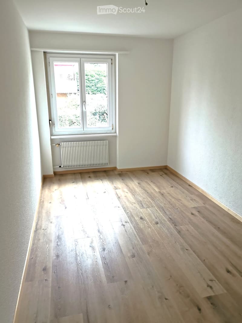 Empty room with light wooden flooring, white walls, and a large window with a radiator below it.