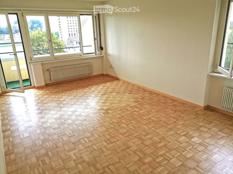 Empty room with wooden herringbone flooring, large windows, radiator, and open door leading to a balcony.