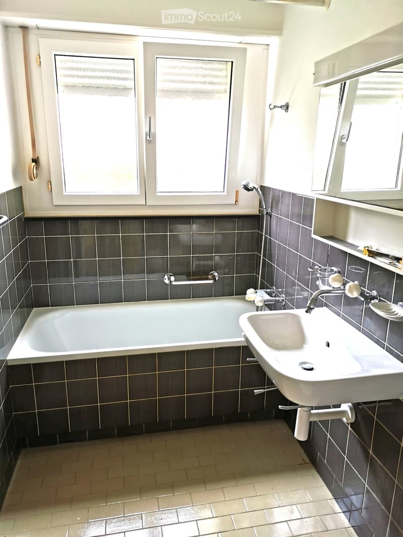Bathroom with a bathtub, sink, and showerhead. Dark tiled walls and light-colored floor tiles.