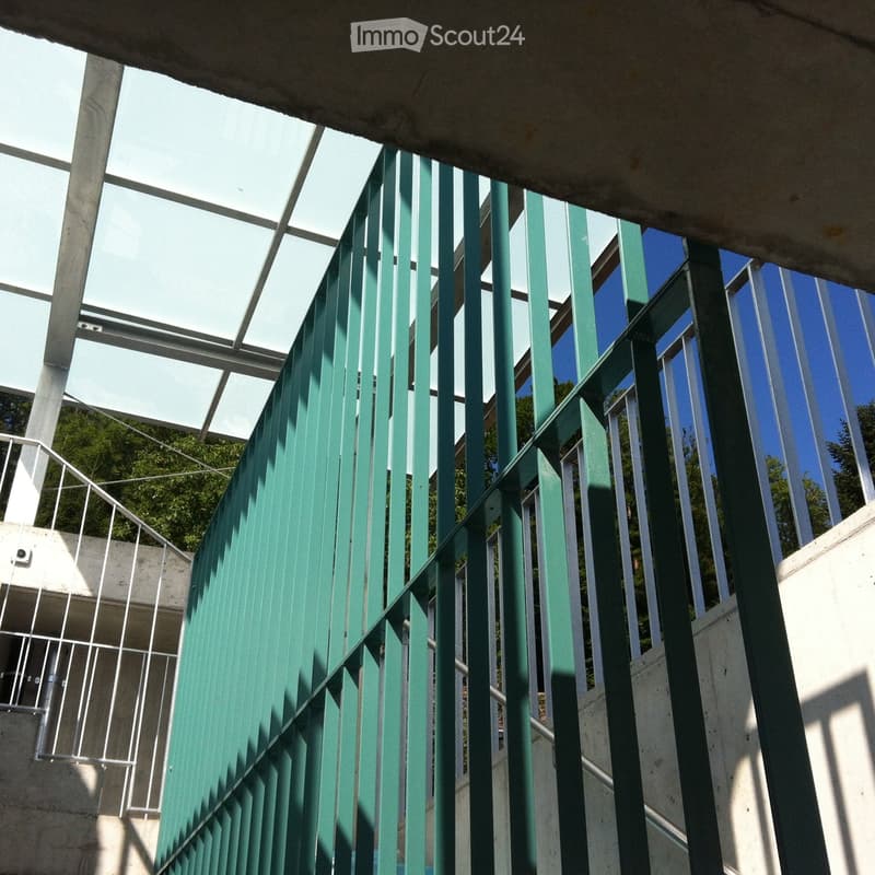 Green metal fence with vertical bars and a glass roof structure, part of a modern building entrance.