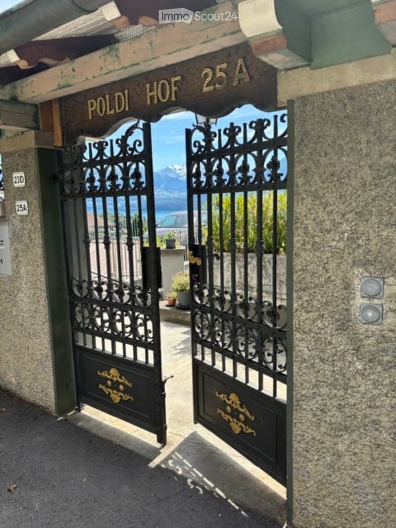 Entrance gate to "Poldi Hof 25A" with ornate black wrought-iron design, featuring gold accents and a view of mountains in the background.