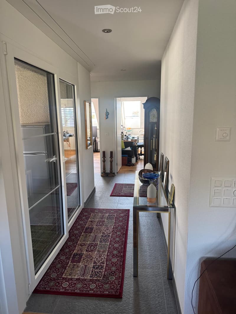 Hallway with tiled floor, red patterned rug, glass table with decorative items, and a view into an adjacent room with a desk and chair.