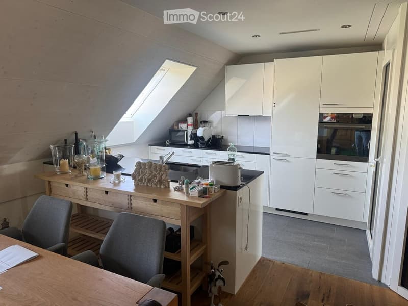 Modern kitchen with white cabinets, stainless steel appliances, and a wooden dining table with chairs. The space features a skylight window and a small workspace area with a sink and various kitchen items.