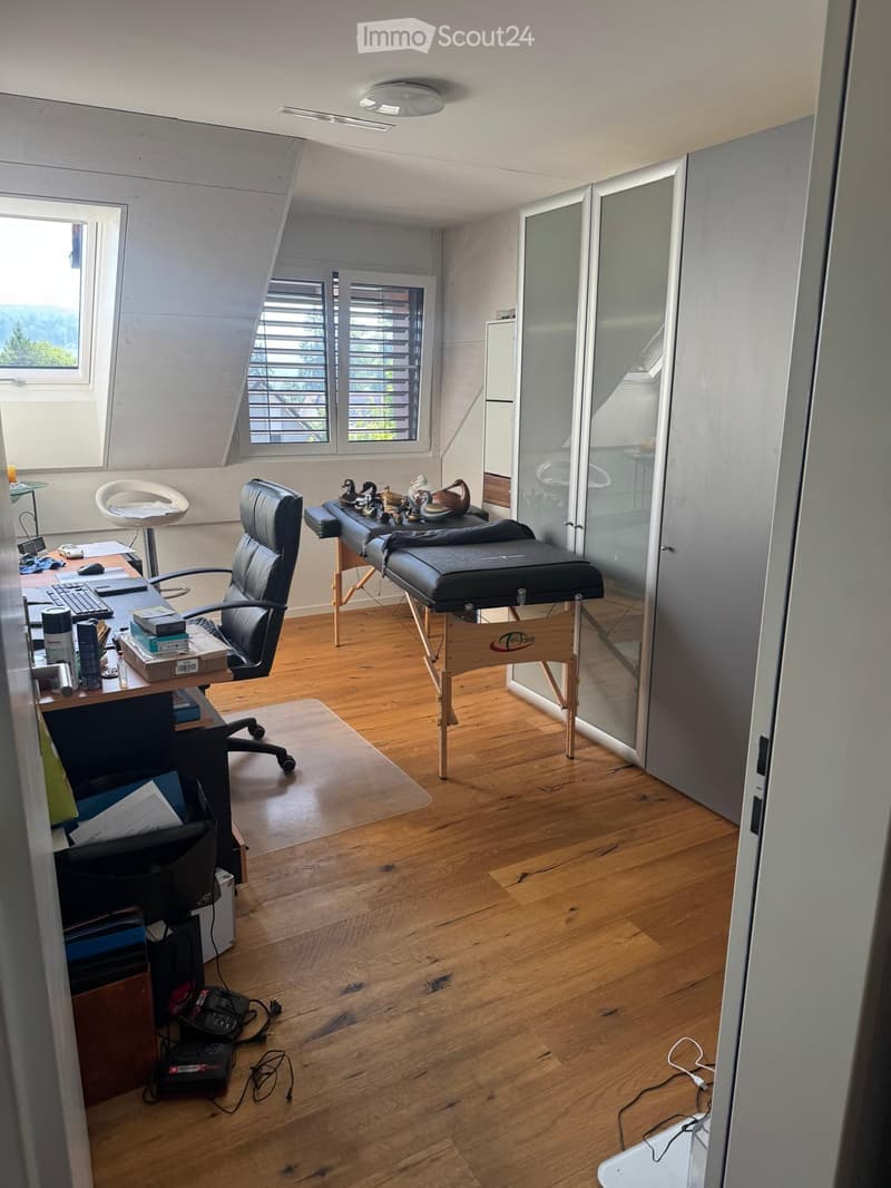 Well-lit room with wooden flooring, a desk with office equipment, a massage table, and decorative items on a small table.
