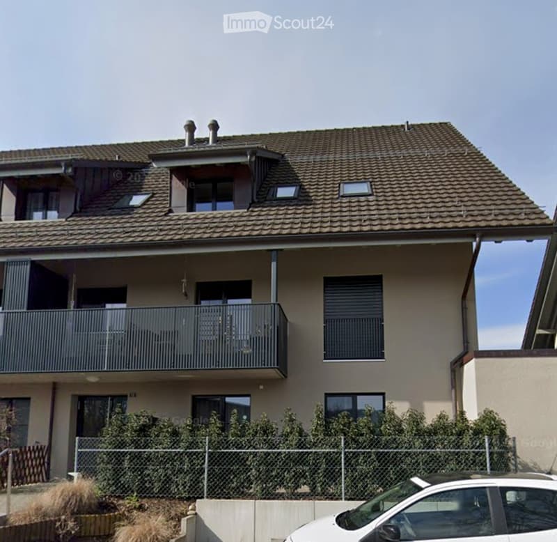 Two-story house with a tiled roof, multiple windows, and a balcony on the upper floor. The house has a light-colored facade and is surrounded by a small garden with a fence.