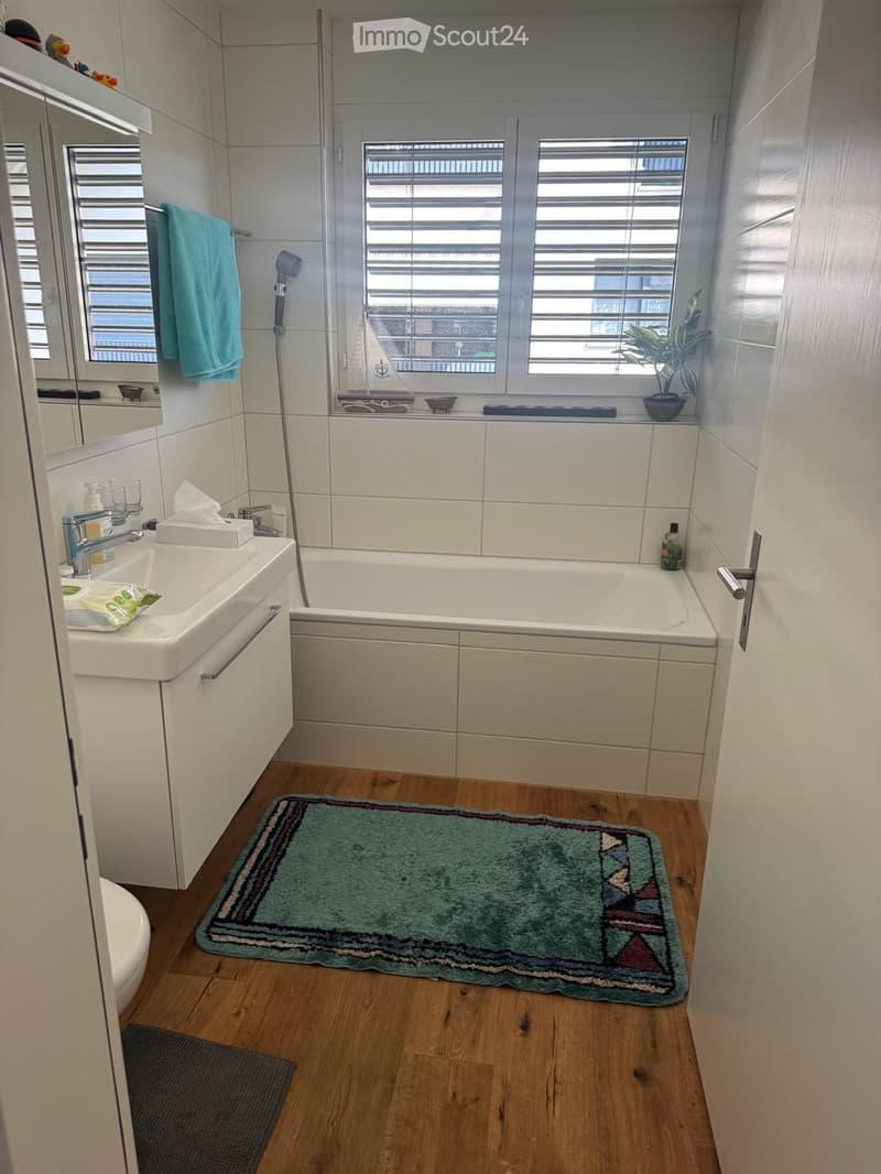 Modern bathroom with a bathtub, sink, mirror, and a green rug on the wooden floor. A towel hangs on the wall, and a small plant sits on the windowsill.