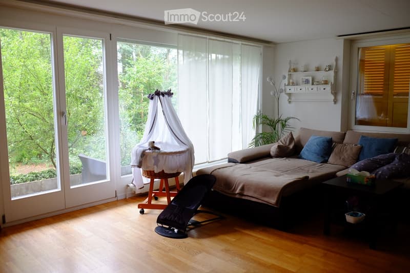 Bright living room with large windows, wooden floor, sofa, baby crib, small table, and decorative shelf.