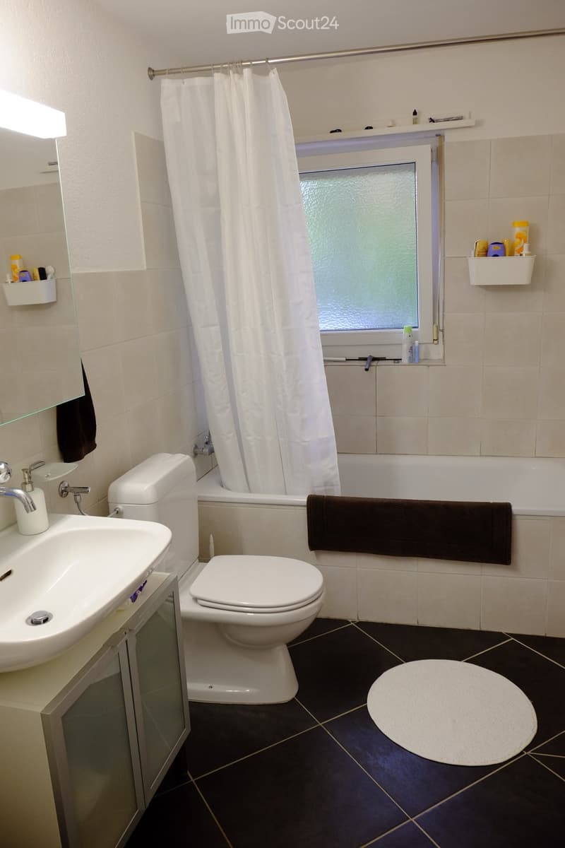 Modern bathroom with a white sink, toilet, bathtub, and shower curtain. Features a dark tiled floor, a round bathroom rug, and a towel hanging on the side of the bathtub.