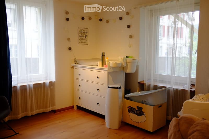 A well-lit bathroom with a bathtub, sink, toilet, and a small cupboard. There is a toy box with a cloud design and a stool near the window. The room has wooden flooring and sheer curtains on the windows.