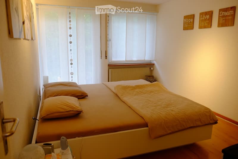 Cozy bedroom with a double bed, pillows, a nightstand with a lamp, and a radiator under the window. The walls are decorated with framed artwork and wooden signs.