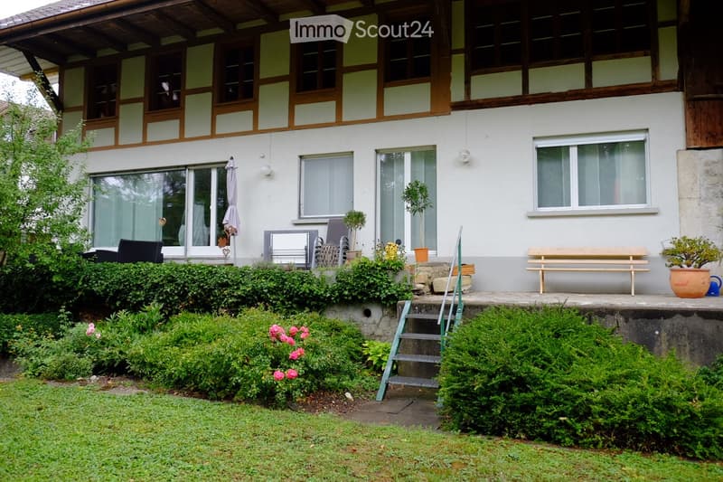 Front garden with well-maintained hedges, flowering shrubs, and a small patio area with a bench, potted plants, and outdoor furniture.