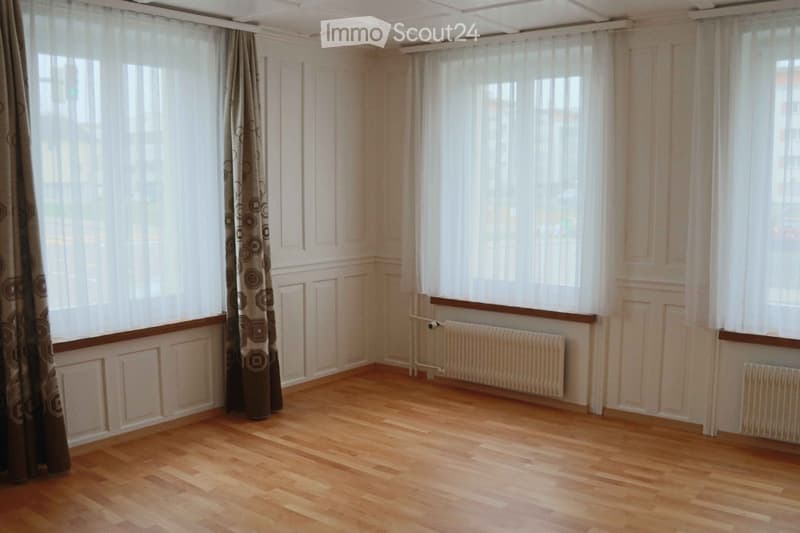 Empty room with wooden flooring, large windows with sheer curtains, and radiators along the walls.