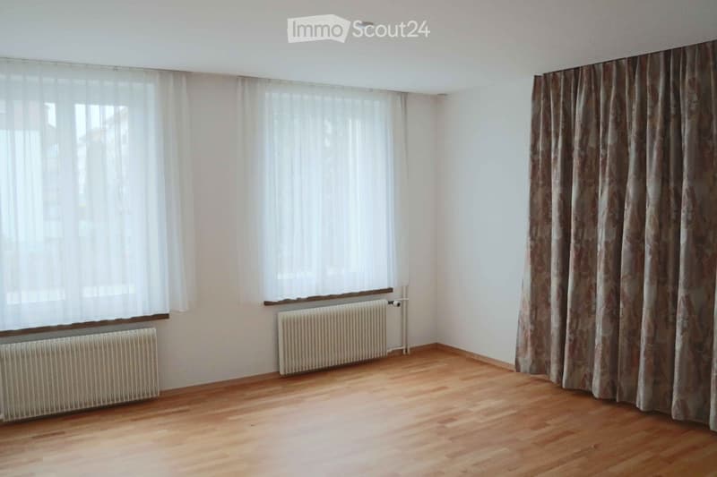 Empty room with wooden flooring, white walls, large windows with curtains, and radiators.