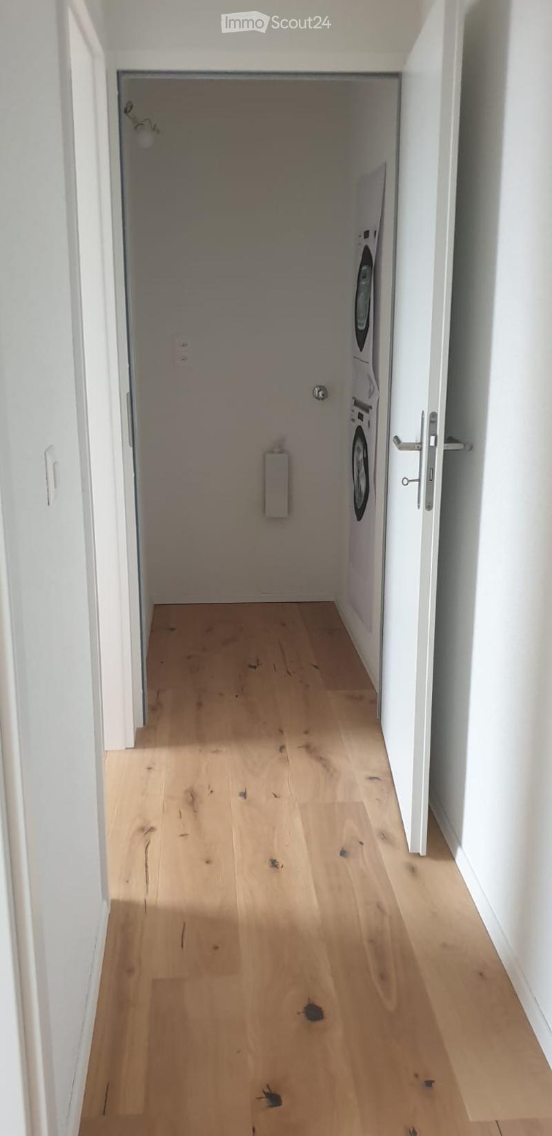 Empty hallway with light wooden flooring and white walls, leading to a room with a washing machine visible at the end.