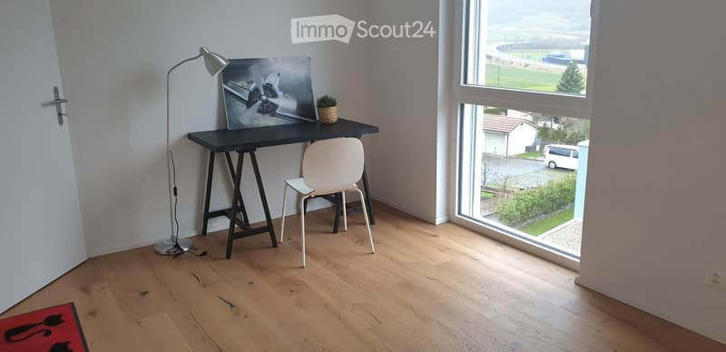 A modern, minimalist room with a wooden floor, large windows offering a view, a black table with a white chair, a floor lamp, a picture frame, and a small plant.