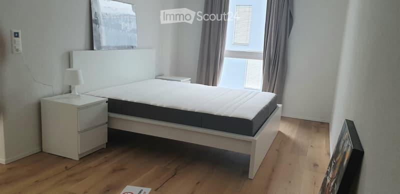 Modern bedroom with a white bed, nightstand, lamp, and picture frame on the wall. The room features wooden flooring and large windows with curtains.