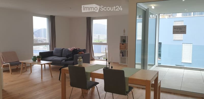 Modern living and dining area with large windows offering a scenic view, featuring a dark blue sofa, wooden coffee table, dining table with chairs, and a small shelf with decorative items.