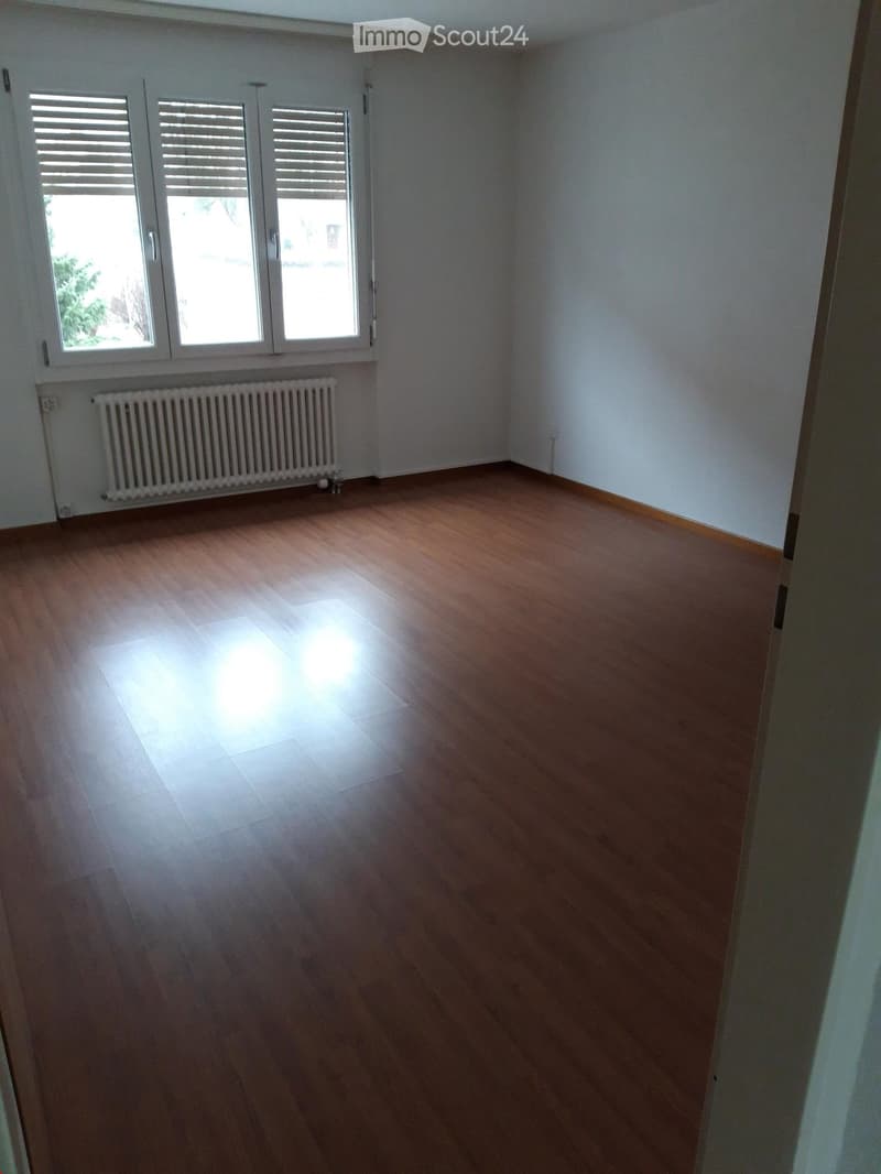 Empty room with wooden flooring, white walls, and a radiator under a window with blinds.