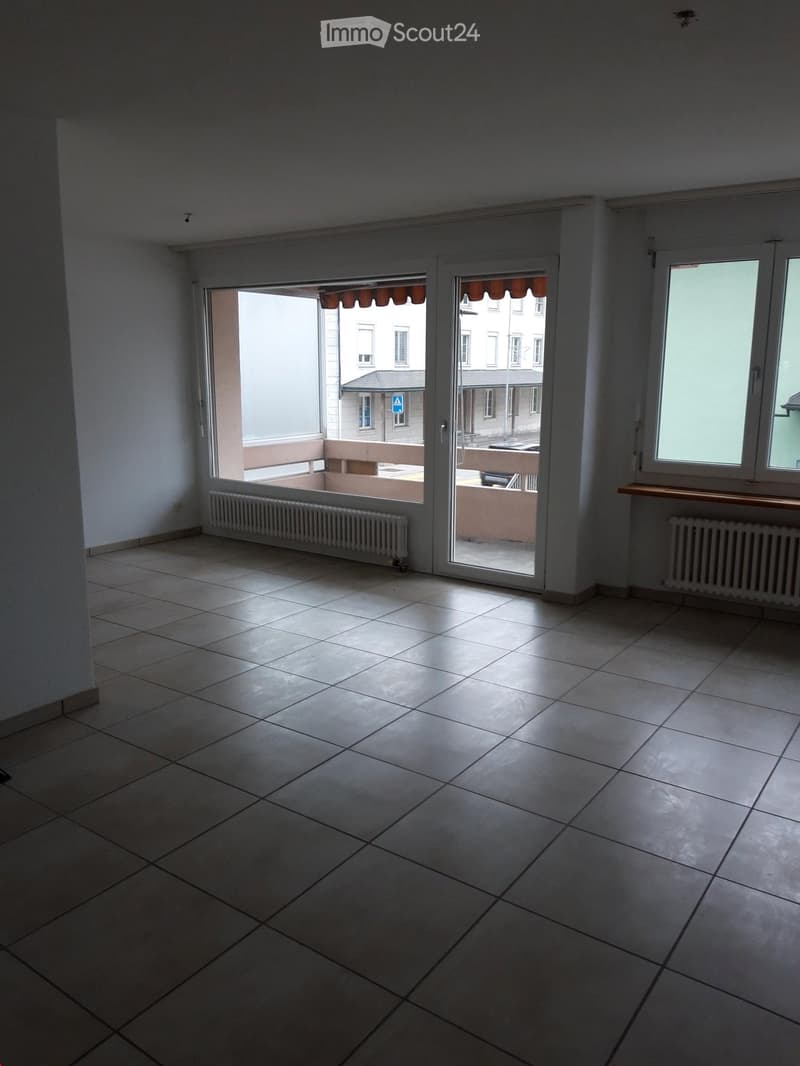 Empty room with tiled floor, white walls, large sliding glass doors leading to a balcony, radiators, and windows.