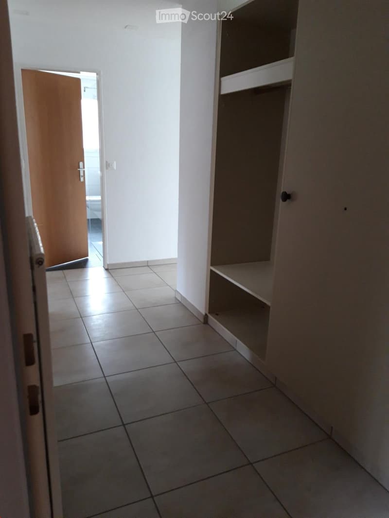 Empty hallway with tiled floor, white walls, and a built-in shelf unit on one side. An open door leads to another room.