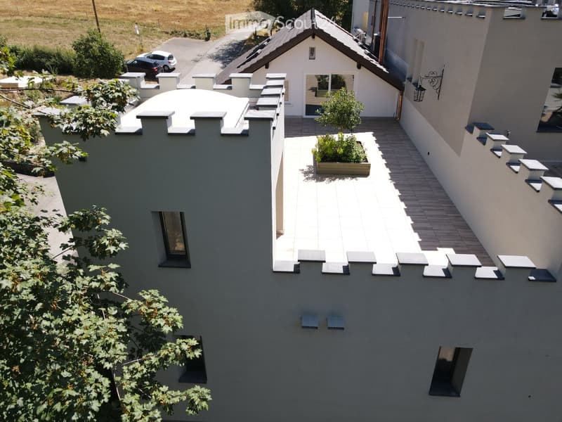Modern two-story building with grey walls, multiple windows, and a small balcony area with a planter and a tree. The roof features a unique design with raised sections and a small dome.